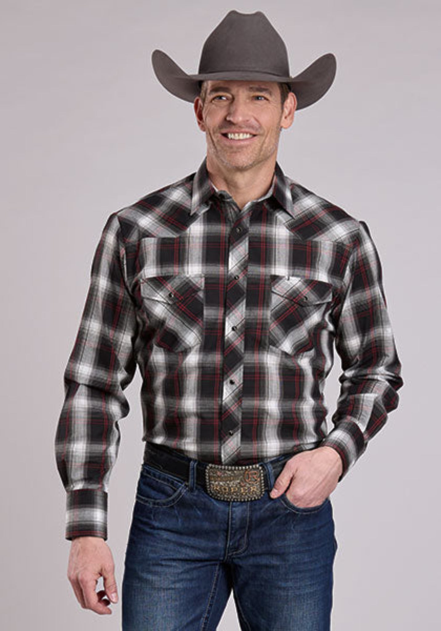 Man wearing a plaid shirt, jeans, and a cowboy hat against a gray background