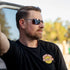 Man wearing sunglasses and a black t-shirt with a logo, sitting in a vehicle.
