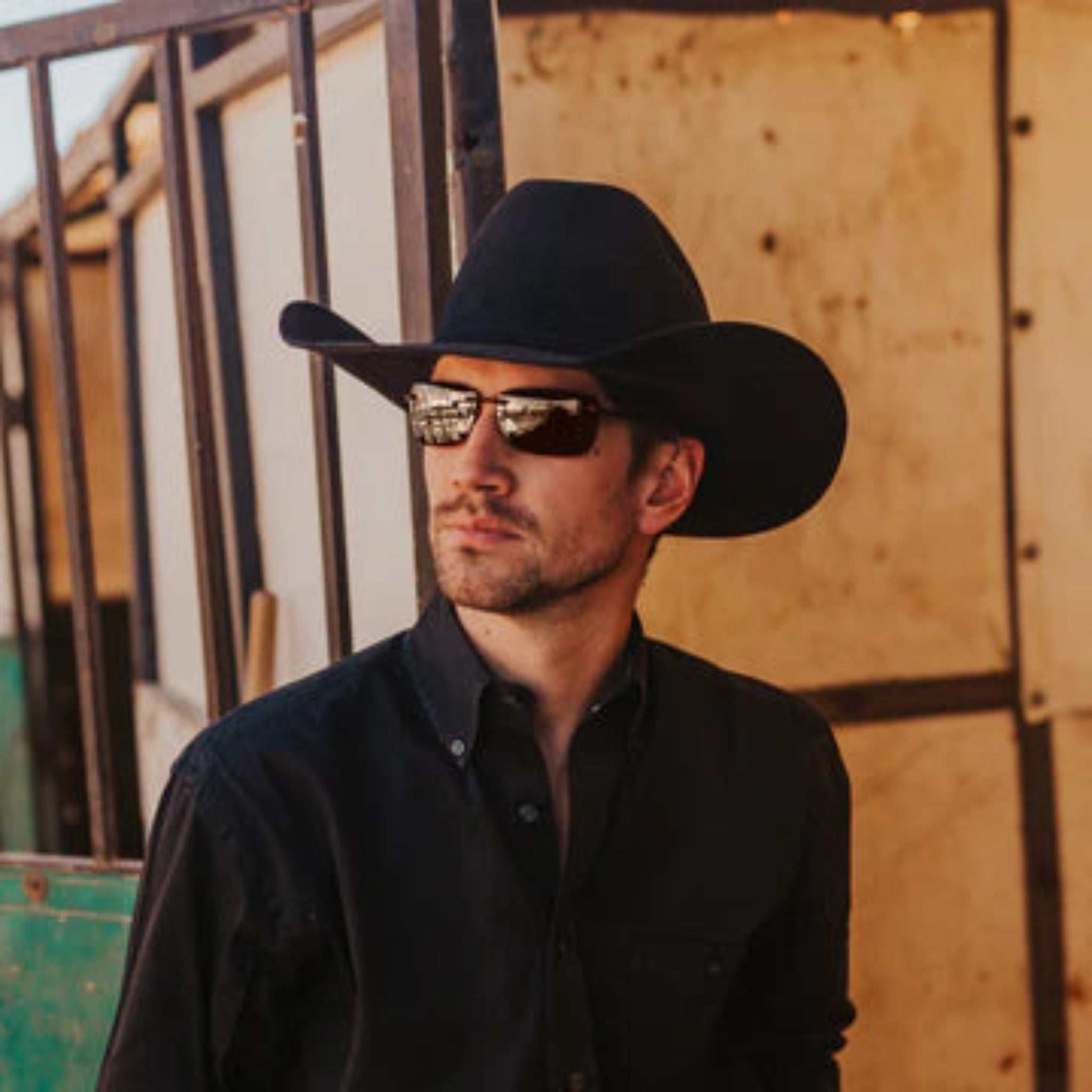 Man wearing a black cowboy hat and sunglasses against a rustic background