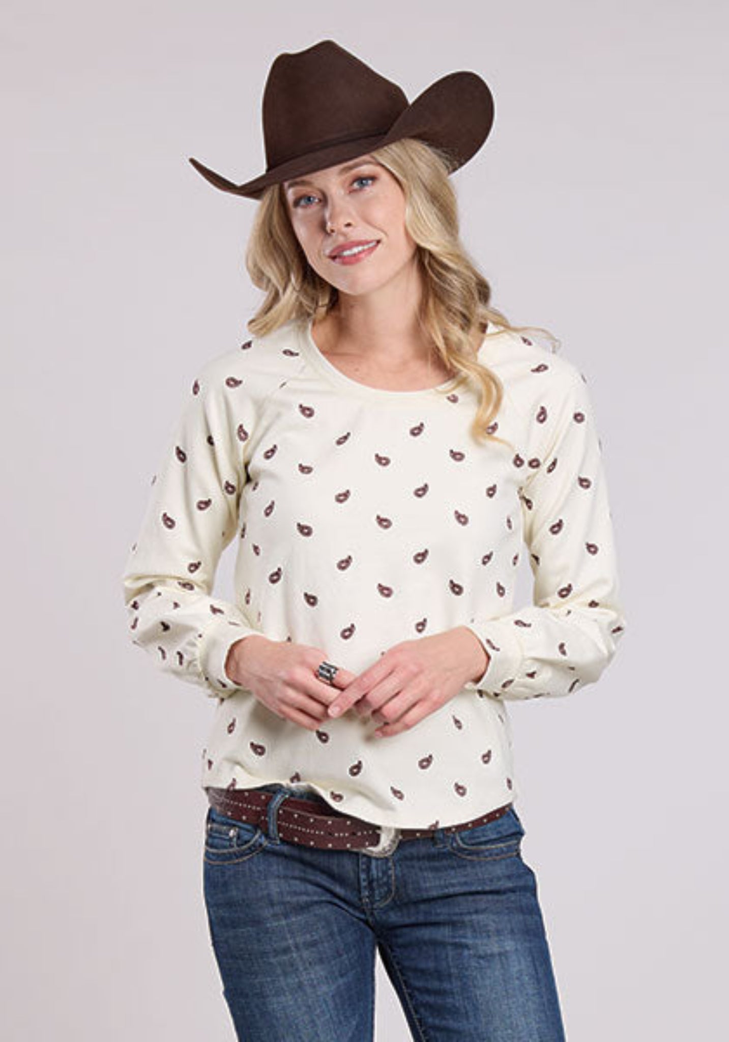 Woman wearing a white long-sleeve shirt with a paisley design, blue jeans, and a brown cowboy hat on a light gray background.