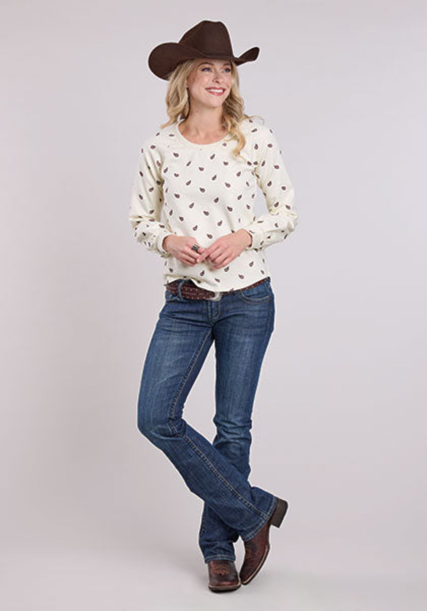 Woman wearing a white patterned shirt, blue jeans, and a brown cowboy hat on a light gray background