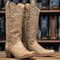 tall western cowboy boots in a distressed tan leather, featuring embossed floral patterns on the shafts and a low stacked heel