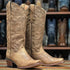 tall western cowboy boots in a distressed tan leather, featuring embossed floral patterns on the shafts and a low stacked heel