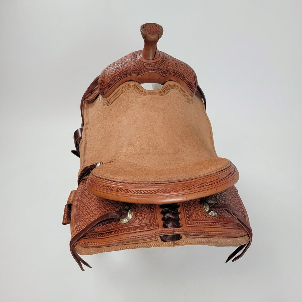A brown leather western saddle with intricate tooling and carved details, photographed indoors against a white fabric backdrop.